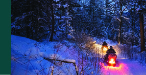 Two snowmobiles on a trail in the woods at dusk.